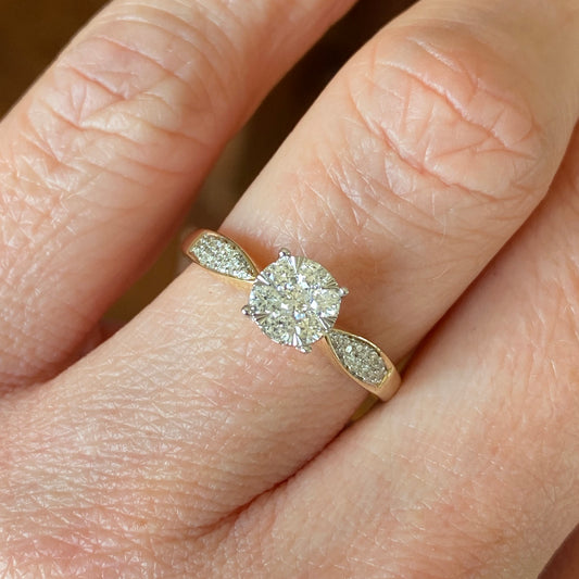 This solitaire cluster diamond engagement ring is a complete classic.  Its simplicity is so romantic.  The Details...  One 9ct gold diamond engagement ring.  Cluster of brilliant cut diamonds.  0.30ct   Colour H.  Clarity SI  Diamond set tapered shoulders with a double row of diamonds.  9ct yellow gold.  Size N